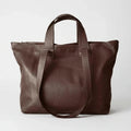 A dark brown leather tote bag with dual top handles and a long shoulder strap, photographed against a clean light gray studio background.