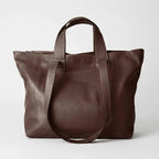 A dark brown leather tote bag with dual top handles and a long shoulder strap, photographed against a clean light gray studio background.