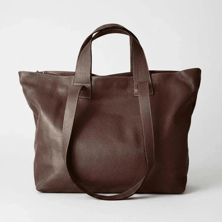 A dark brown leather tote bag with dual top handles and a long shoulder strap, photographed against a clean light gray studio background.
