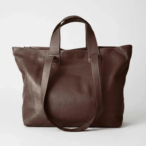 A dark brown leather tote bag with dual top handles and a long shoulder strap, photographed against a clean light gray studio background.