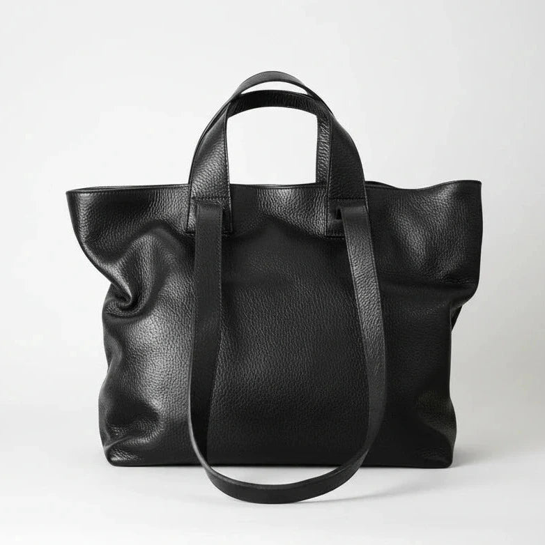 A black pebbled leather tote bag with dual top handles and a long shoulder strap, photographed against a clean light gray studio background.