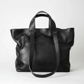 A black pebbled leather tote bag with dual top handles and a long shoulder strap, photographed against a clean light gray studio background.