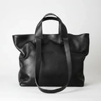 A black pebbled leather tote bag with dual top handles and a long shoulder strap, photographed against a clean light gray studio background.