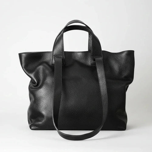 A black pebbled leather tote bag with dual top handles and a long shoulder strap, photographed against a clean light gray studio background.