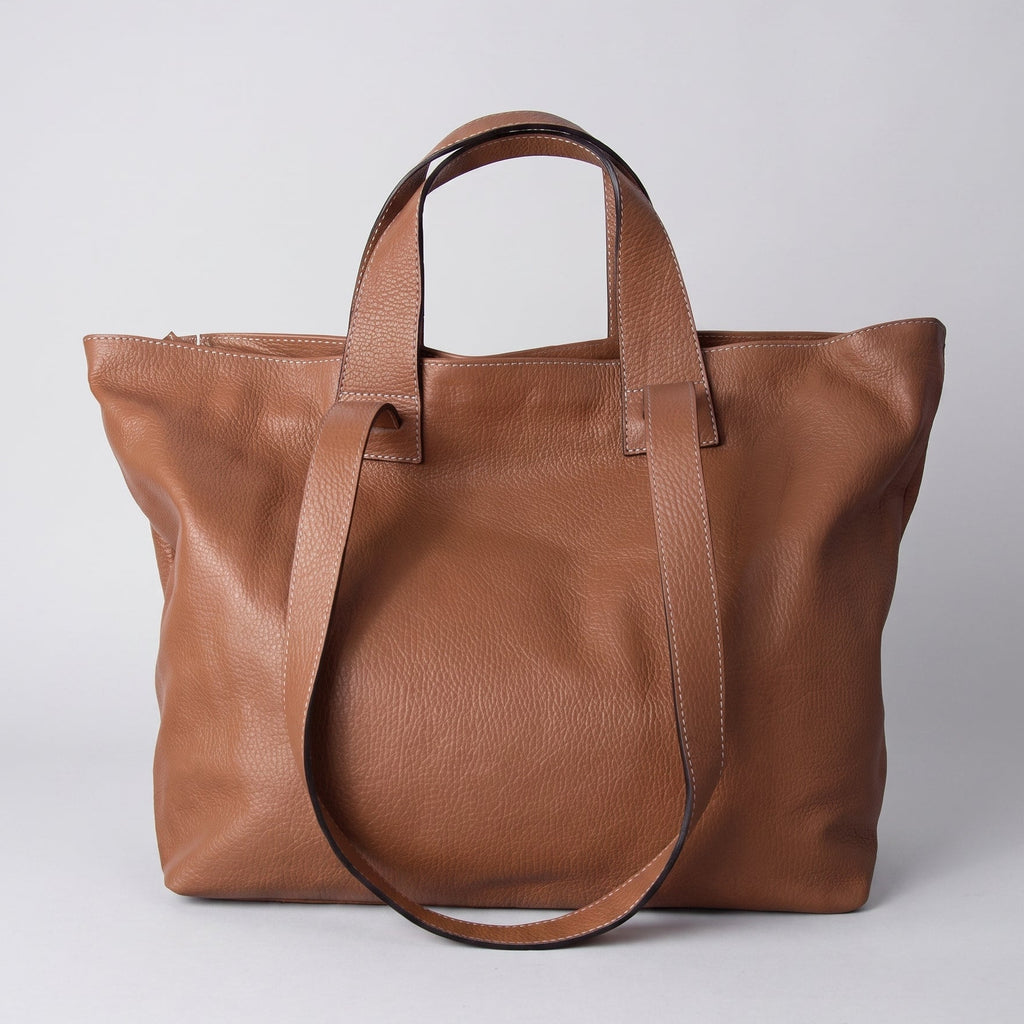 A medium brown leather tote bag with dual top handles and a detachable shoulder strap, photographed against a neutral light gray background.
