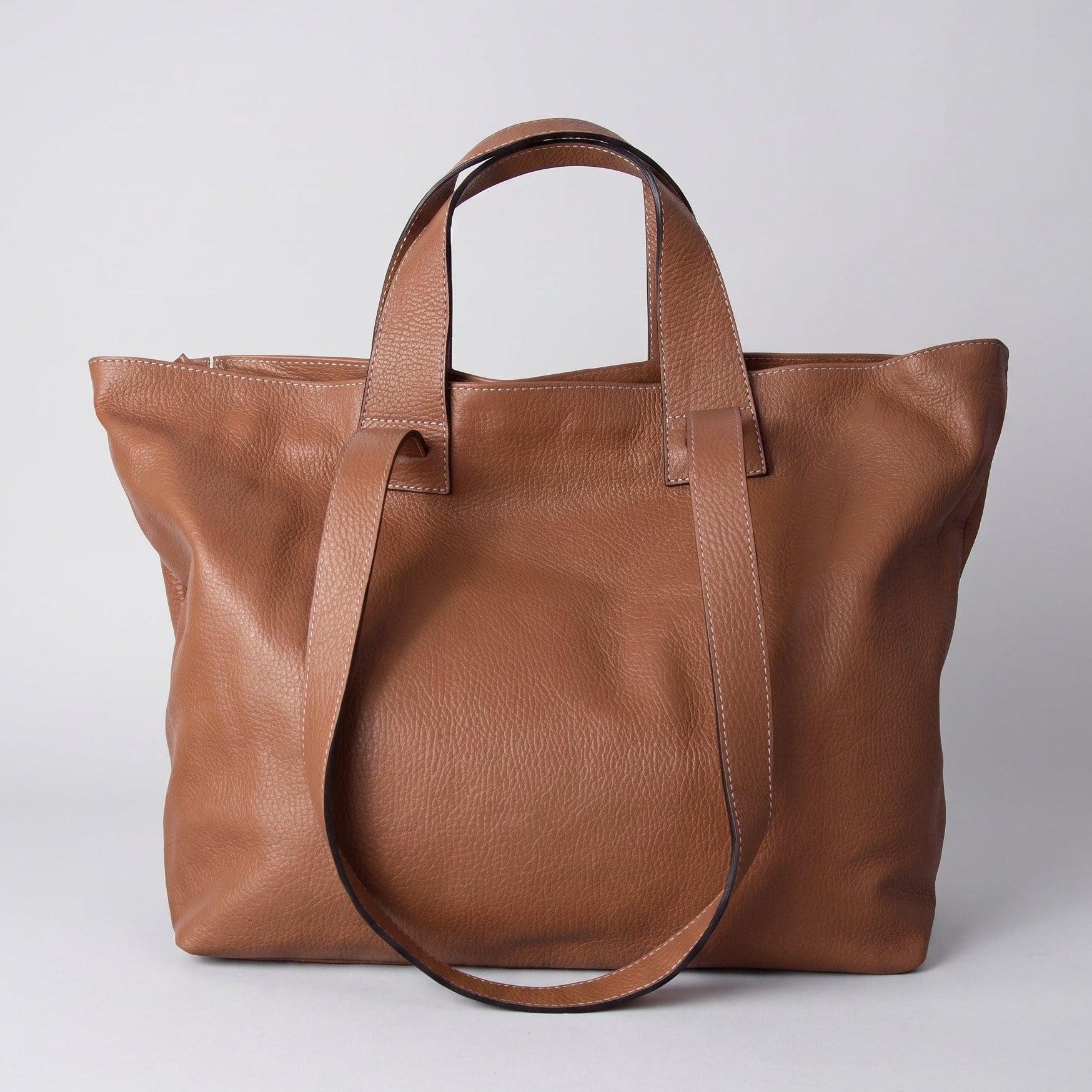 A medium brown leather tote bag with dual top handles and a detachable shoulder strap, photographed against a neutral light gray background.