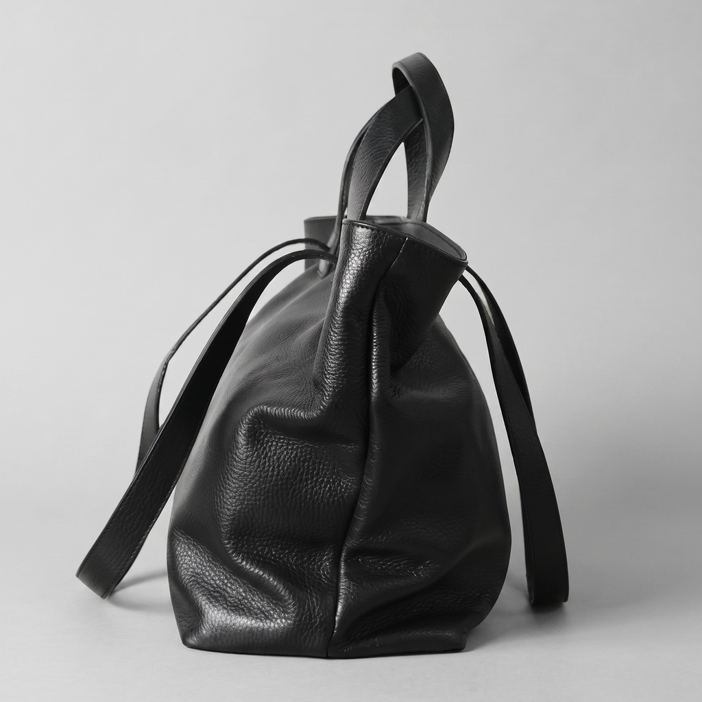 A black pebbled leather bucket bag with dual top handles and a long shoulder strap, photographed against a neutral light gray background.
