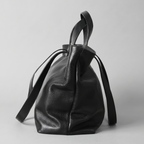 A black pebbled leather bucket bag with dual top handles and a long shoulder strap, photographed against a neutral light gray background.