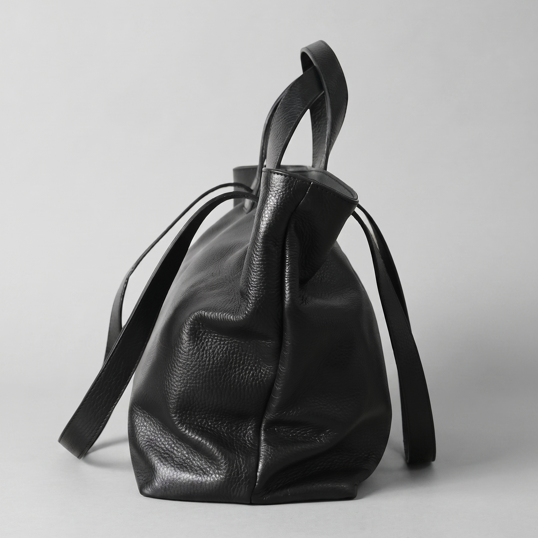 A black pebbled leather bucket bag with dual top handles and a long shoulder strap, photographed against a neutral light gray background.