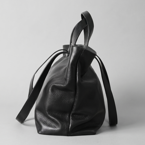 A black pebbled leather bucket bag with dual top handles and a long shoulder strap, photographed against a neutral light gray background.