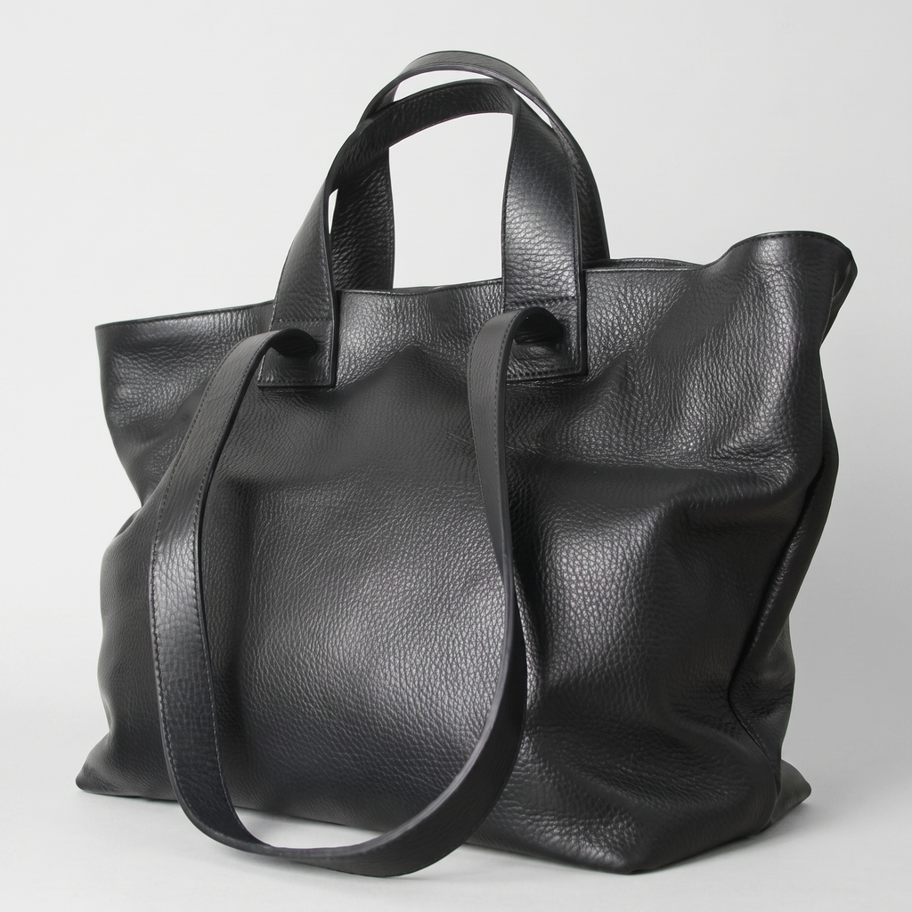 A black pebbled leather tote bag with dual top handles and a long shoulder strap, photographed on a light gray studio background.