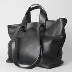 A black pebbled leather tote bag with dual top handles and a long shoulder strap, photographed on a light gray studio background.