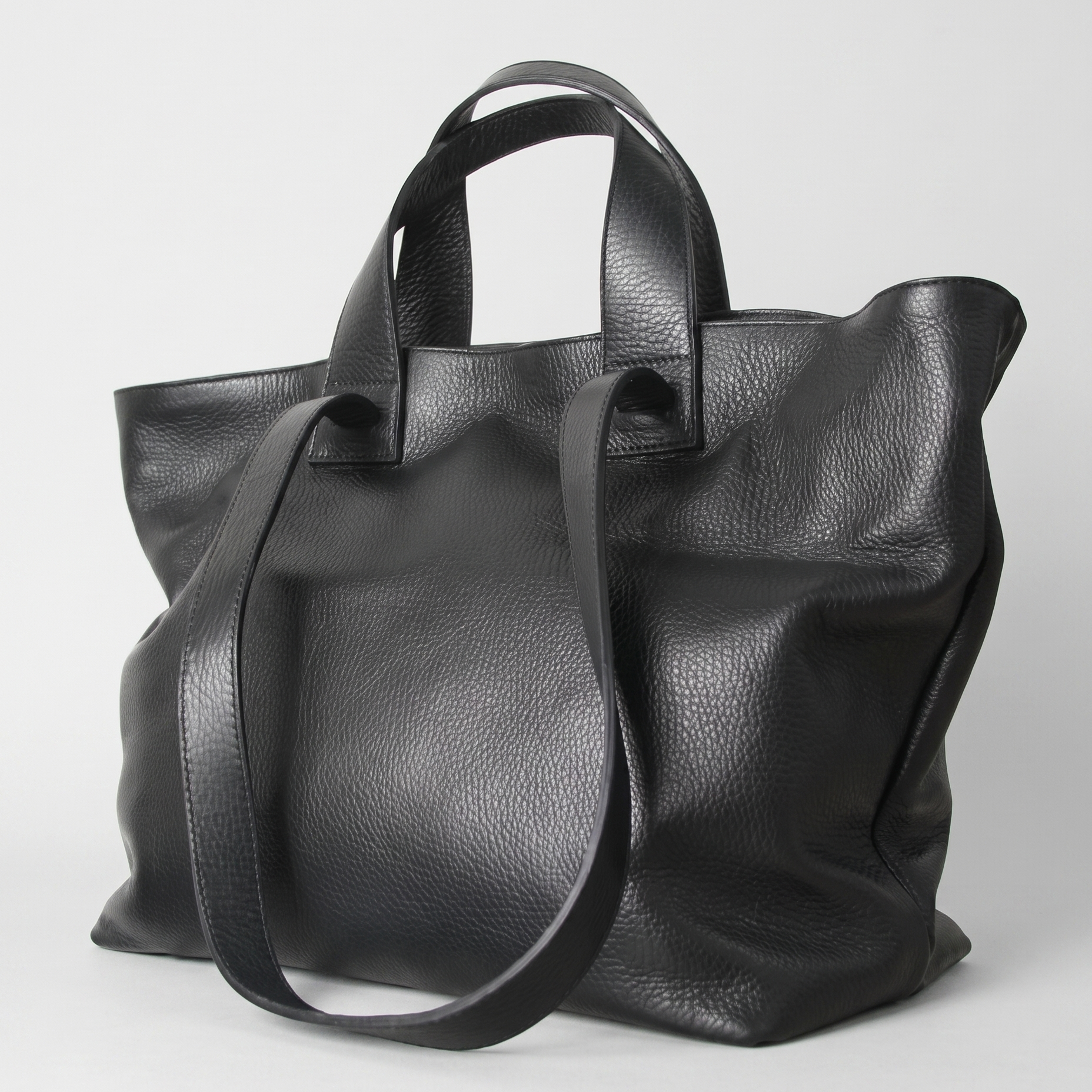 A black pebbled leather tote bag with dual top handles and a long shoulder strap, photographed on a light gray studio background.